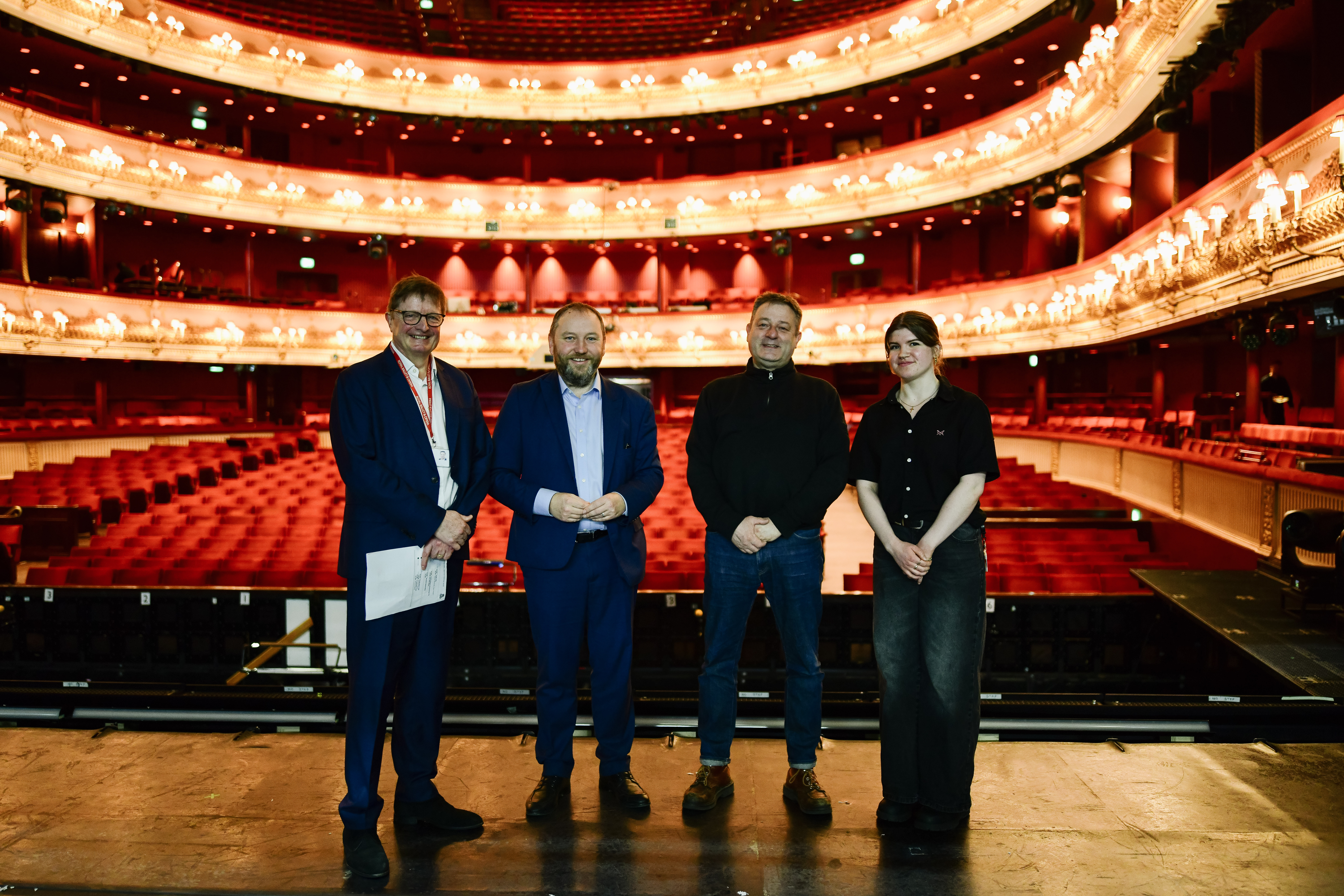 Four people stand together on stage, posing for a photo. Behind them is the grand Royal Opera House auditorium.