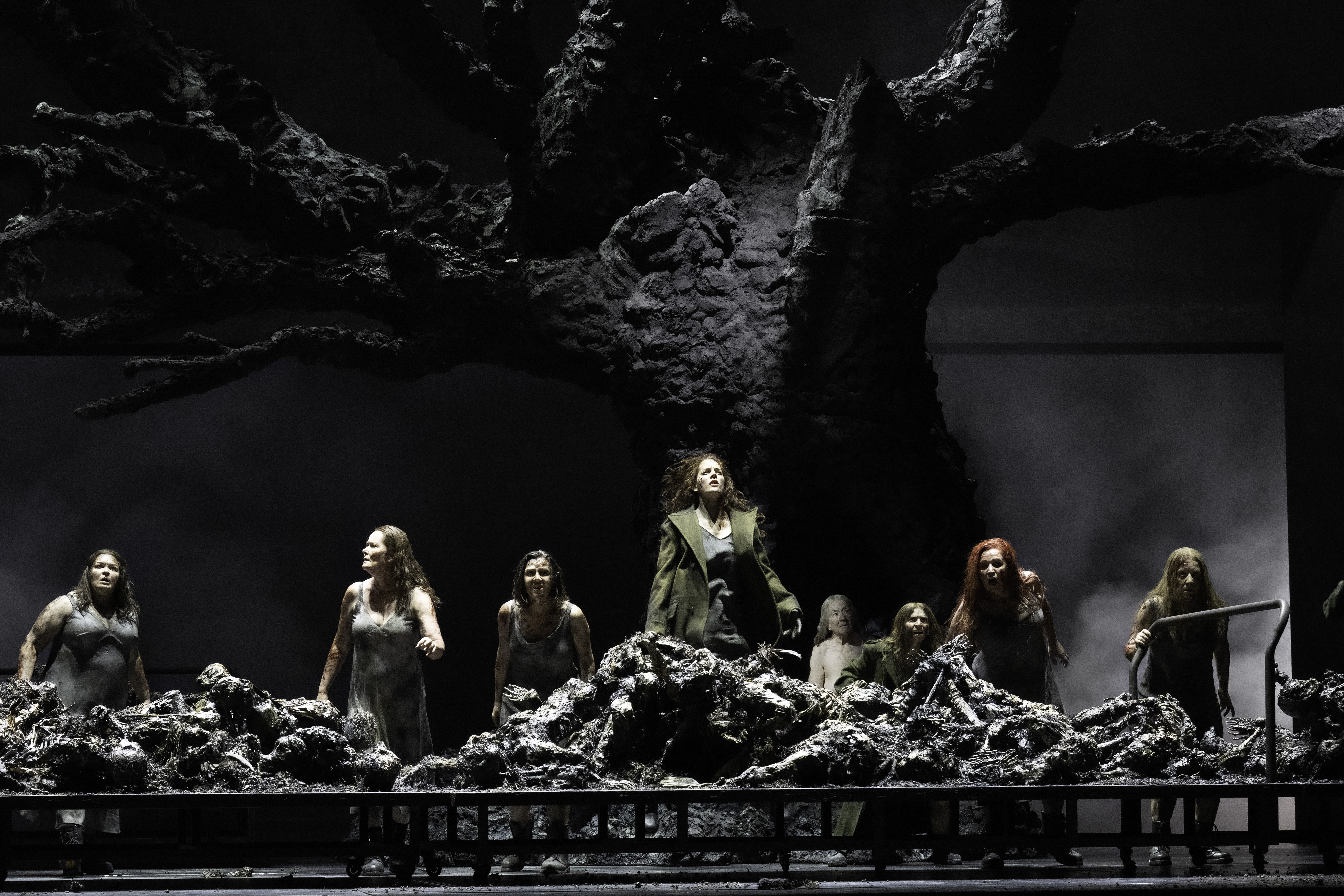 A group of performers stand in front of a blackened tree. They stand, looking shocked, amongst the charred remains of warriors. (3 of 6)