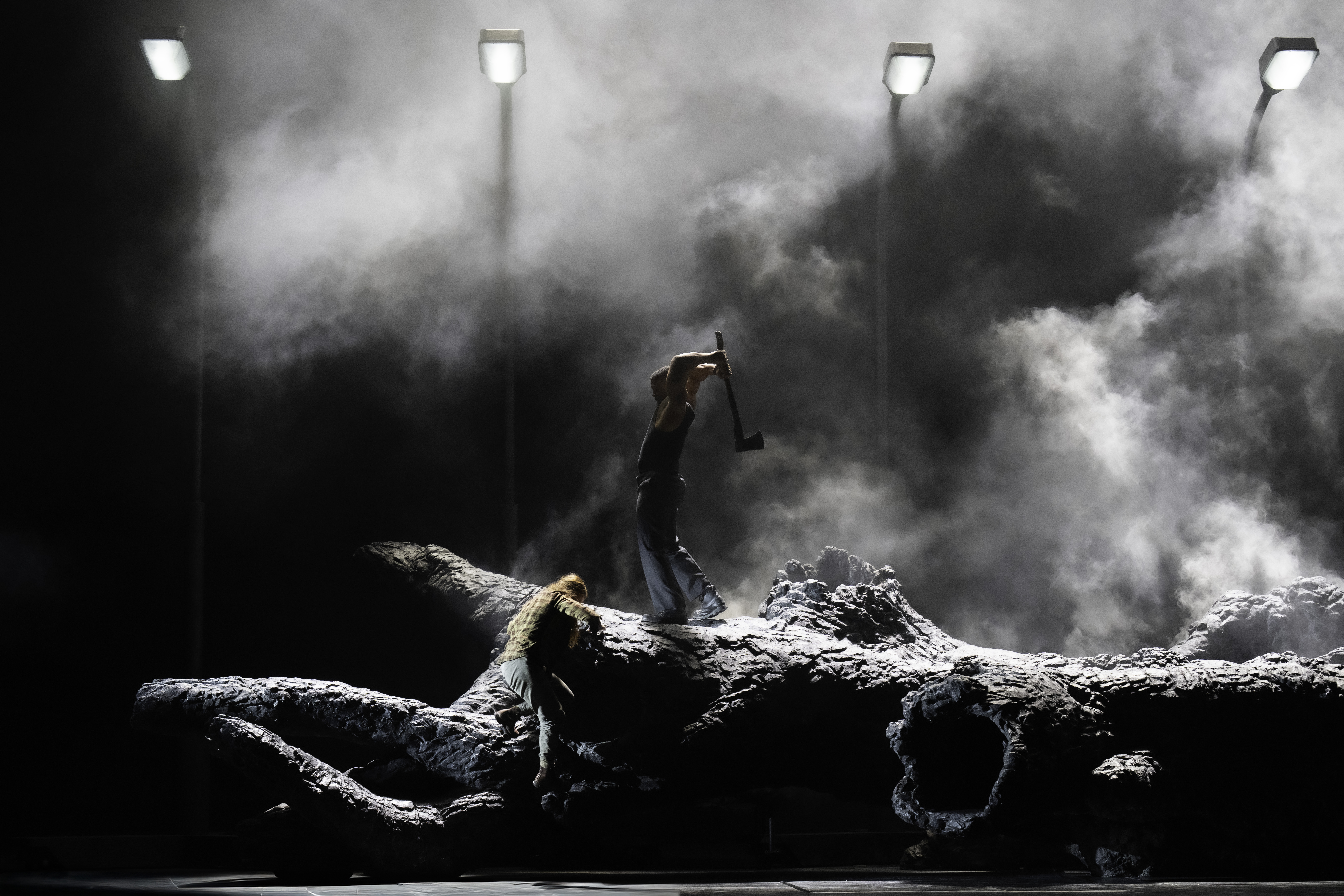 A man stands atop a felled tree - he swings an axe above his head. Below him a woman holds onto the trunk of the tree. Behind them street lamps appear from the smoke and darkness of the stage. (4 of 6)