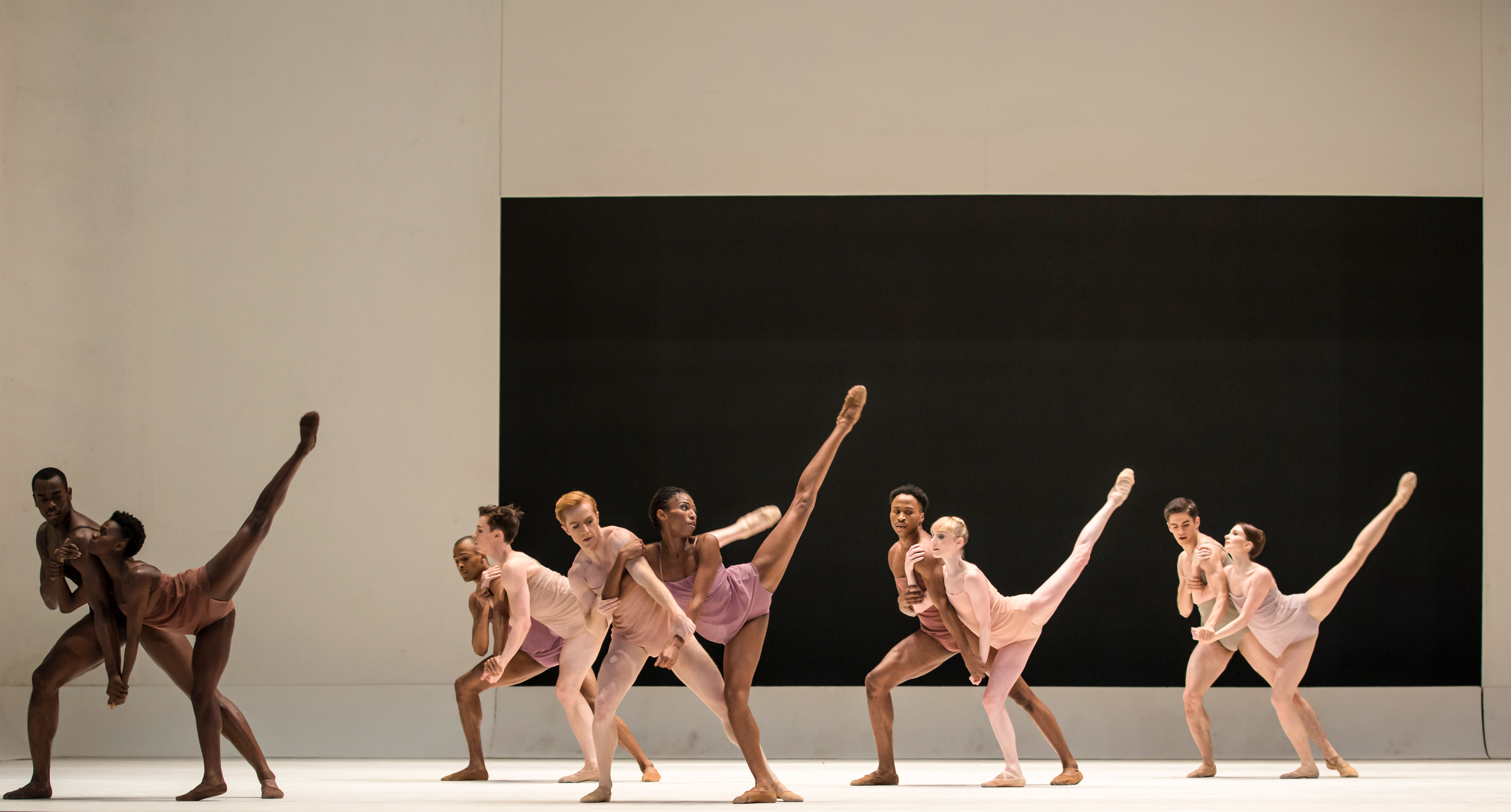 A group of dancers stand on stage, they wear skin coloured costumes. The stage is bare except for a black rectangle on the backdrop. The female dancers hold a their legs in the air. (2 of 10)