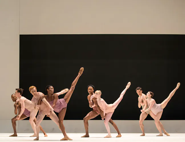 A group of dancers stand on stage, they wear skin coloured costumes. The stage is bare except for a black rectangle on the backdrop. The female dancers hold a their legs in the air. (2 of 10)