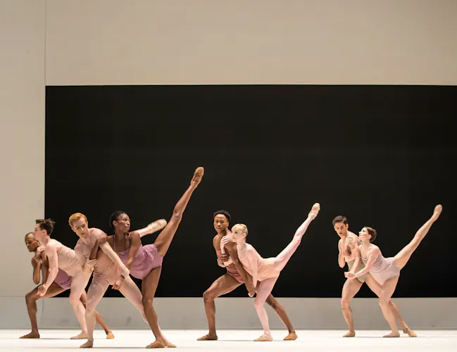 A group of dancers stand on stage, they wear skin coloured costumes. The stage is bare except for a black rectangle on the backdrop. The female dancers hold a their legs in the air. (5 of 8)