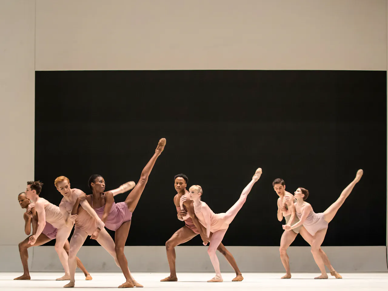 A group of dancers stand on stage, they wear skin coloured costumes. The stage is bare except for a black rectangle on the backdrop. The female dancers hold a their legs in the air.