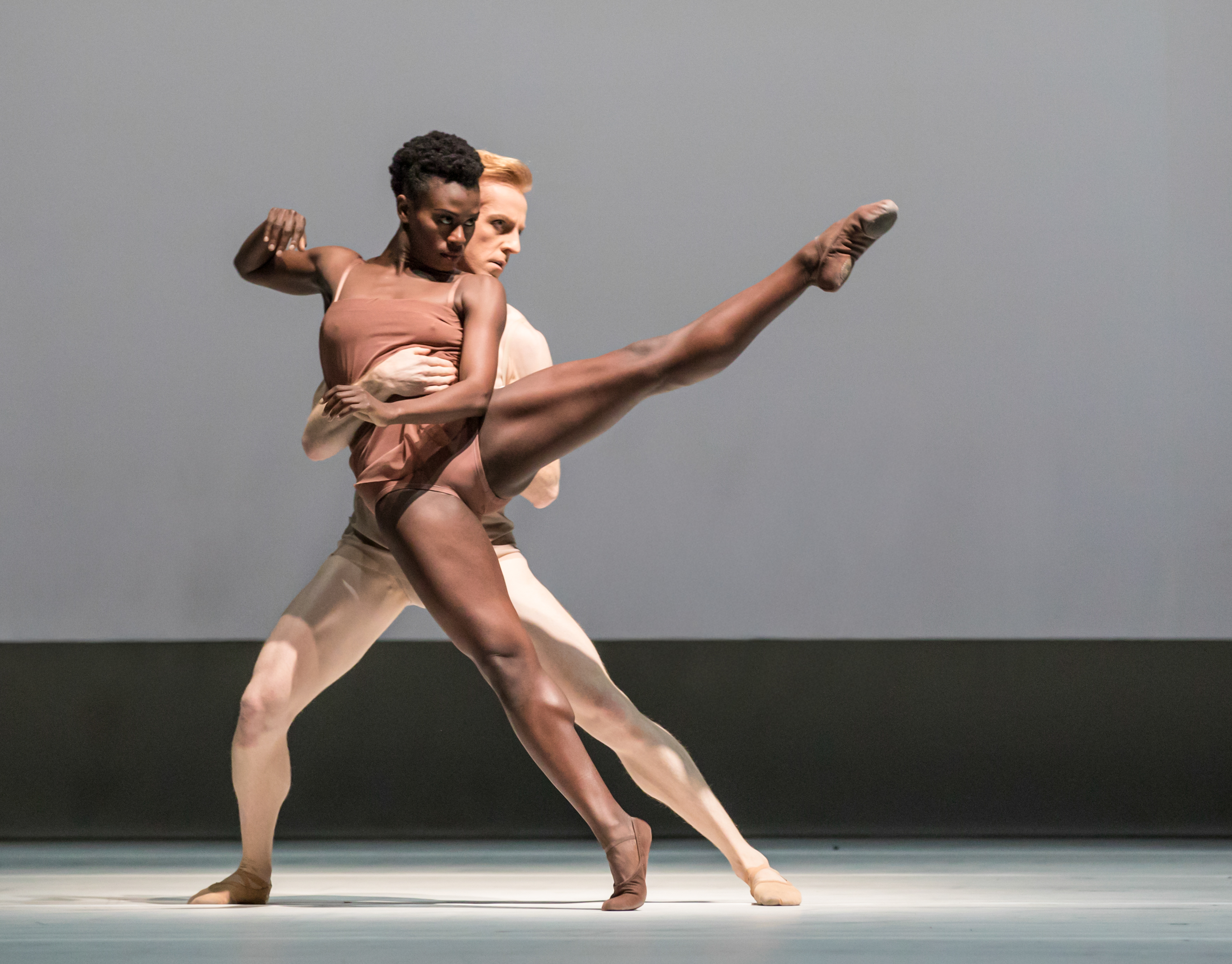 Two dancers perform on stage. They wear skin coloured outifts. The female dancer holds a pose with her left leg outstretched, the male dancer supporting her from behind. (6 of 8)