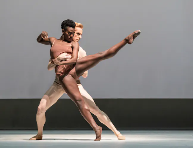 Two dancers perform on stage. They wear skin coloured outifts. The female dancer holds a pose with her left leg outstretched, the male dancer supporting her from behind. (6 of 8)