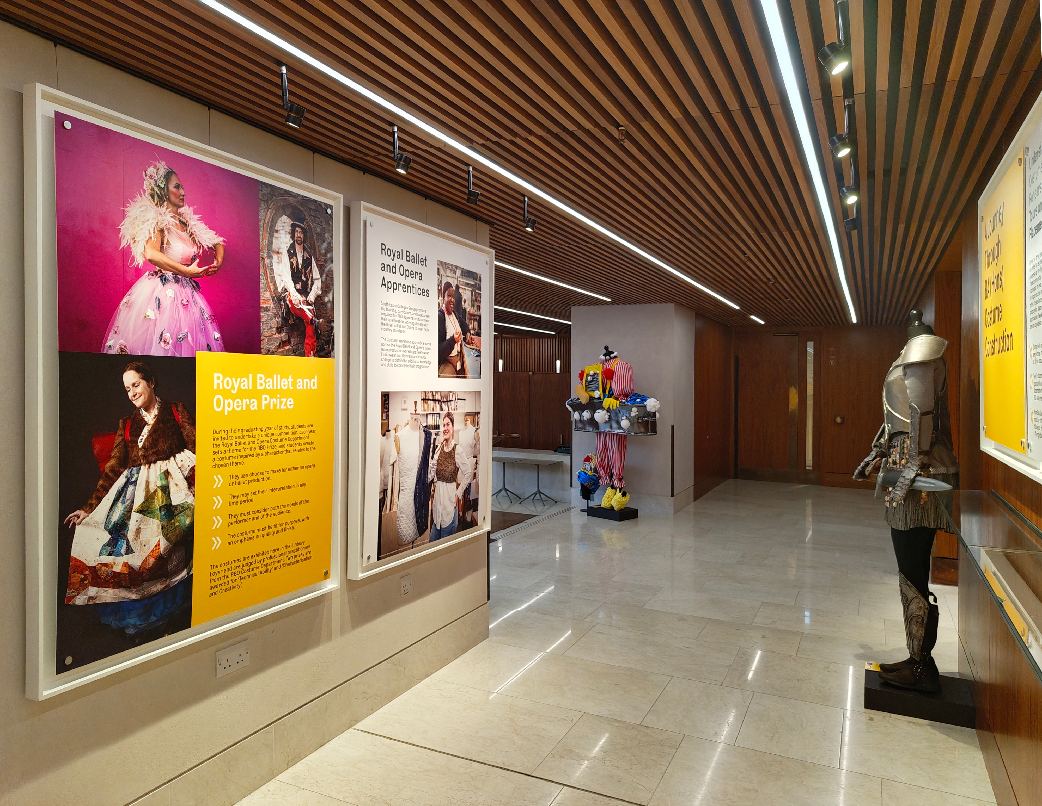 A modern gallery space with wooden ceiling panels, large framed photos and text about the Royal Ballet and Opera Prize on the walls, a mannequin in a colourful dress, and a suit of armour displayed on the right.