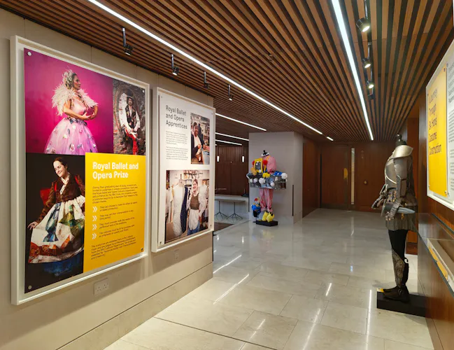 A modern gallery space with wooden ceiling panels, large framed photos and text about the Royal Ballet and Opera Prize on the walls, a mannequin in a colourful dress, and a suit of armour displayed on the right.