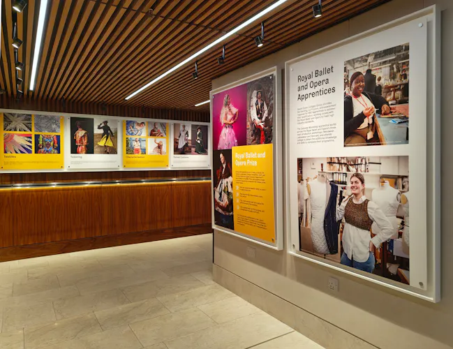 A modern gallery space with wooden ceiling panels, large framed photos and text about the Royal Ballet and Opera Prize on the walls.