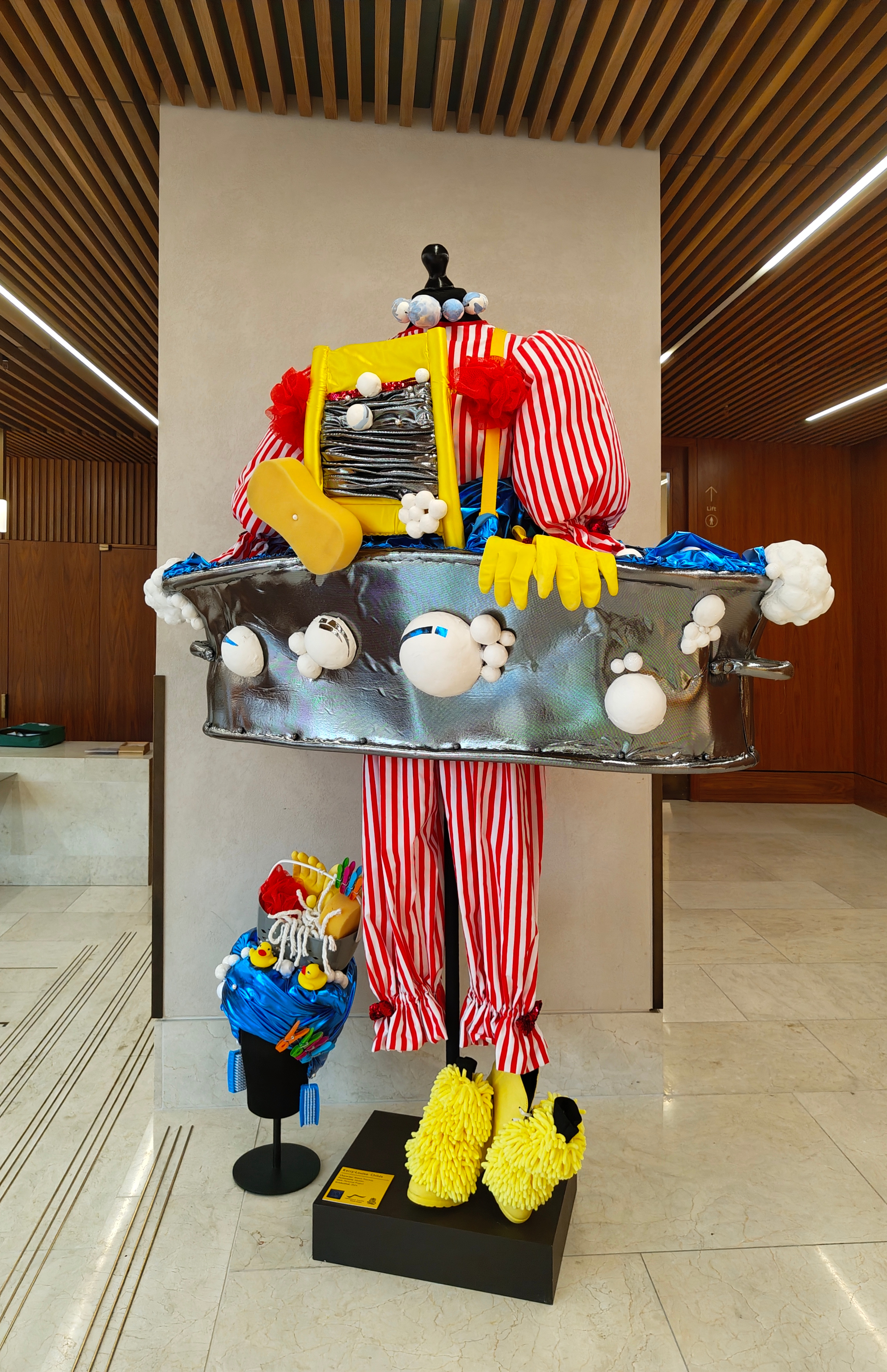 A whimsical sculpture with red-and-white striped pants, oversized yellow shoes, and a metallic tub-like body decorated with plush, colourful objects, displayed indoors on a marble floor with a wood-panelled background.