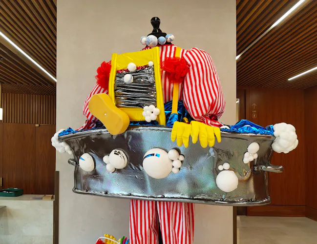 A whimsical sculpture with red-and-white striped pants, oversized yellow shoes, and a metallic tub-like body decorated with plush, colourful objects, displayed indoors on a marble floor with a wood-panelled background.