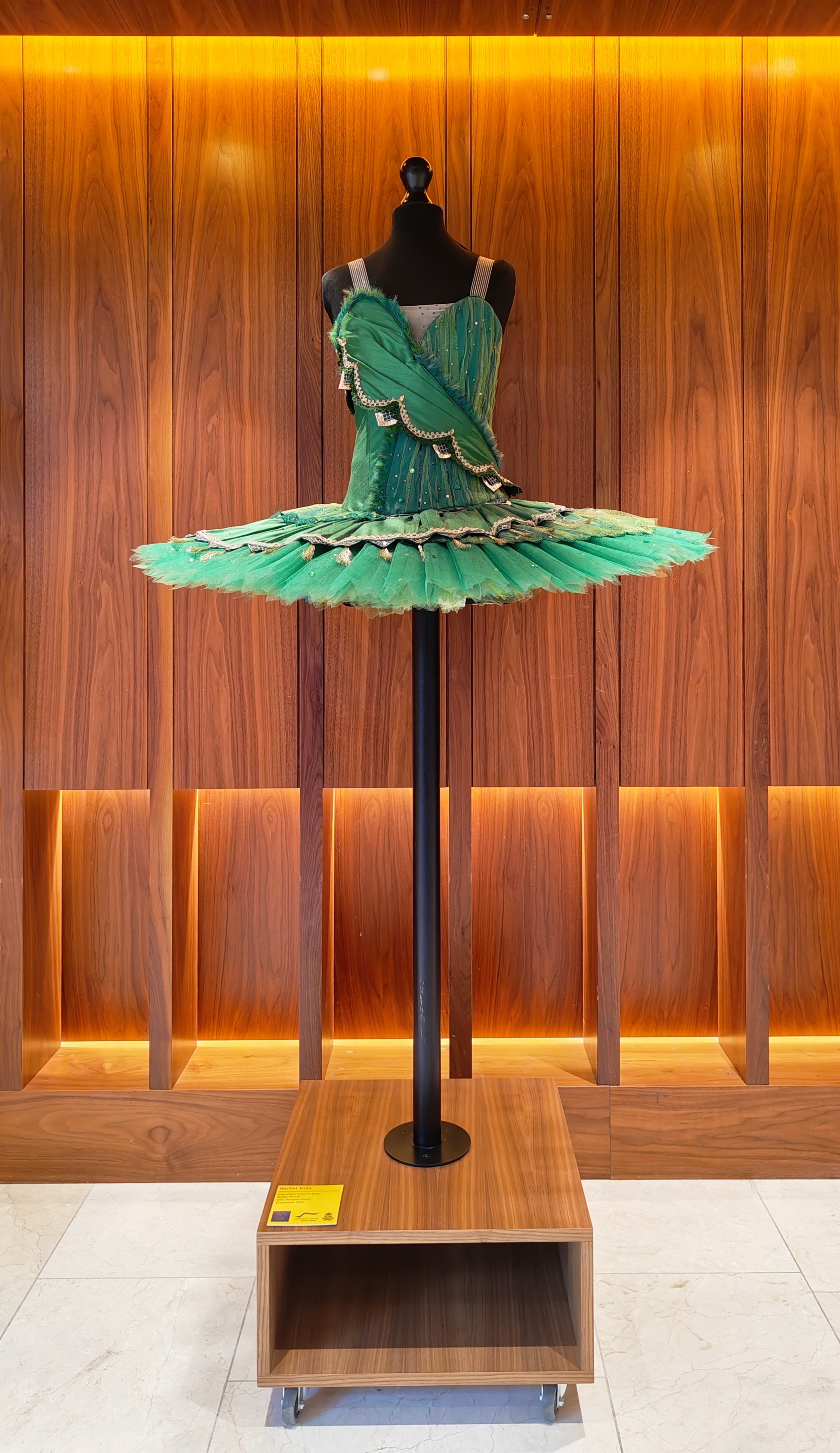 A green ballet tutu with gold and black accents displayed on a black mannequin against a wood-panelled wall with warm lighting, on a wooden pedestal.