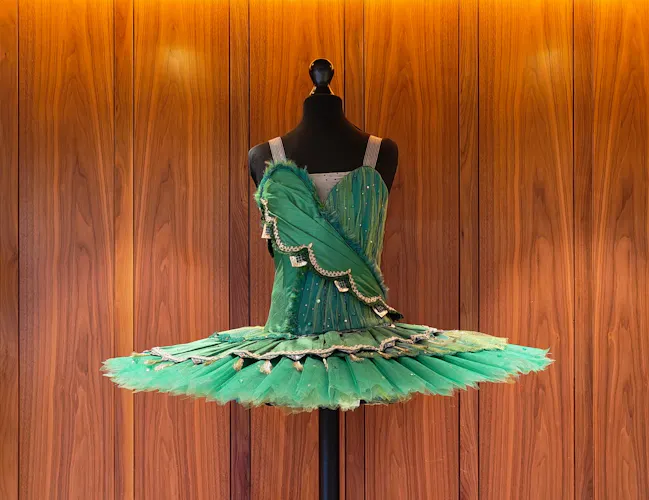 A green ballet tutu with gold and black accents displayed on a black mannequin against a wood-panelled wall with warm lighting, on a wooden pedestal.