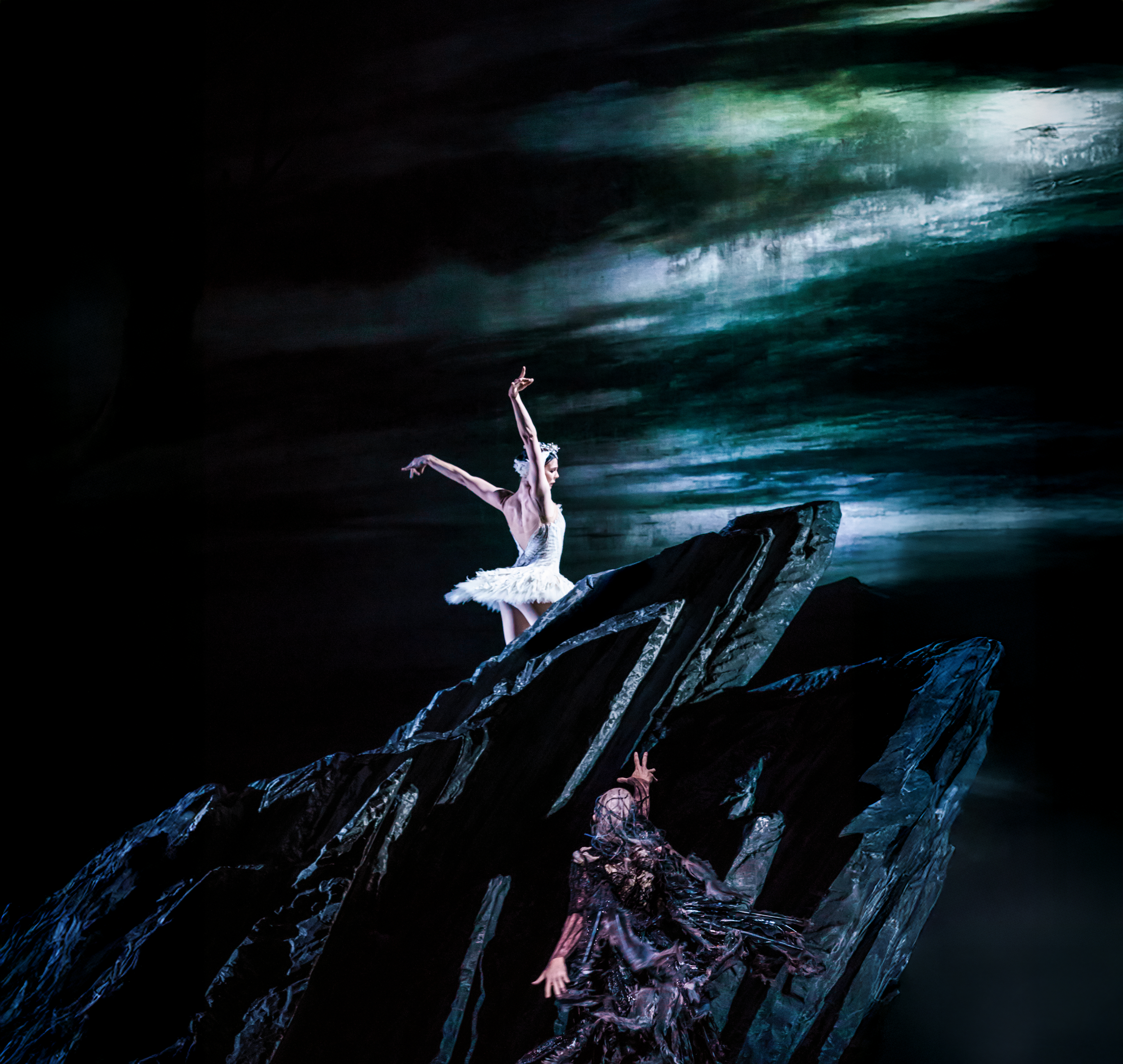 The Royal Ballet dancer Natalia Osipova wears a white tutu and feathered headdress. She stands atop a black rock. Below the rock, the villainous Von Rothbard reaches up towards her, he wears a black feathered cape. Behind them both is a painted backdrop of a dark stormy sky, with a lake in the foreground They are performing in The Royal Ballet's production of Swan Lake.