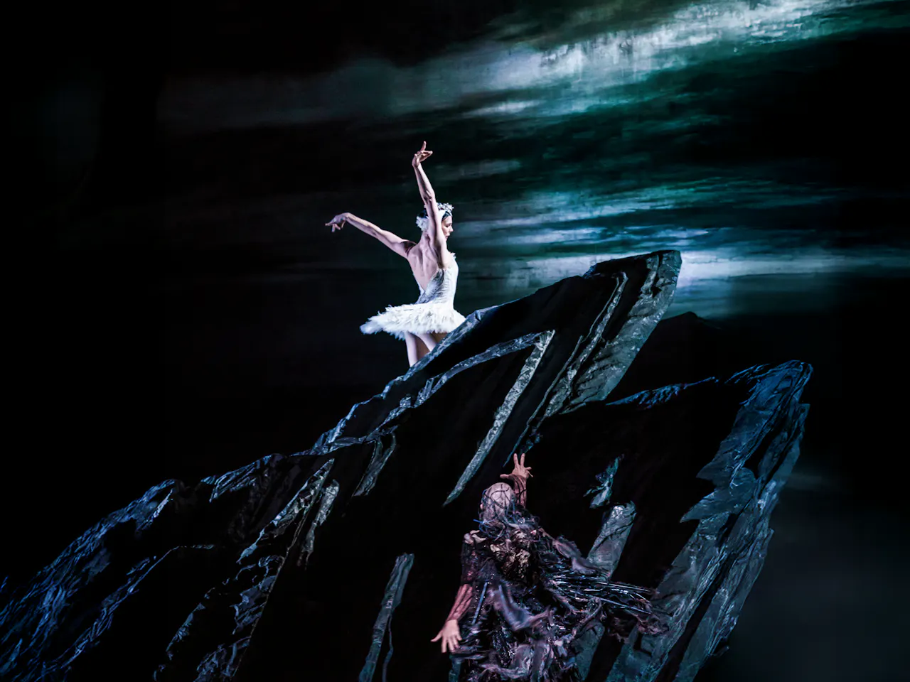 The Royal Ballet dancer Natalia Osipova wears a white tutu and feathered headdress. She stands atop a black rock. Below the rock, the villainous Von Rothbard reaches up towards her, he wears a black feathered cape. Behind them both is a painted backdrop of a dark stormy sky, with a lake in the foreground They are performing in The Royal Ballet's production of Swan Lake.