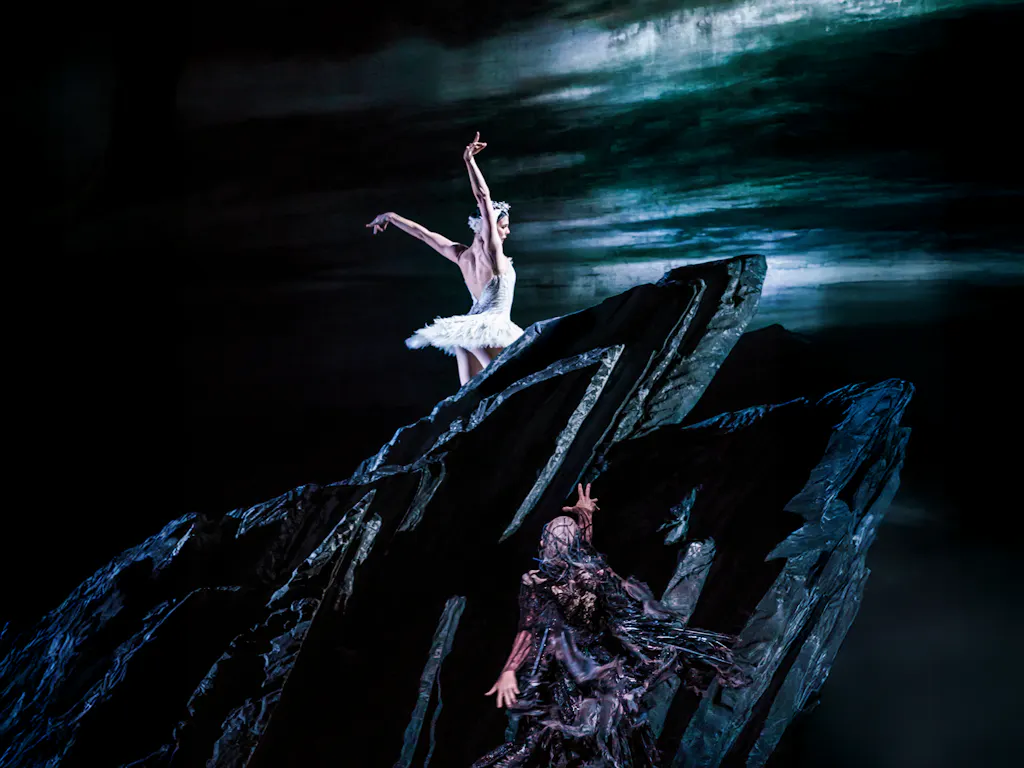 The Royal Ballet dancer Natalia Osipova wears a white tutu and feathered headdress. She stands atop a black rock. Below the rock, the villainous Von Rothbard reaches up towards her, he wears a black feathered cape. Behind them both is a painted backdrop of a dark stormy sky, with a lake in the foreground They are performing in The Royal Ballet's production of Swan Lake.
