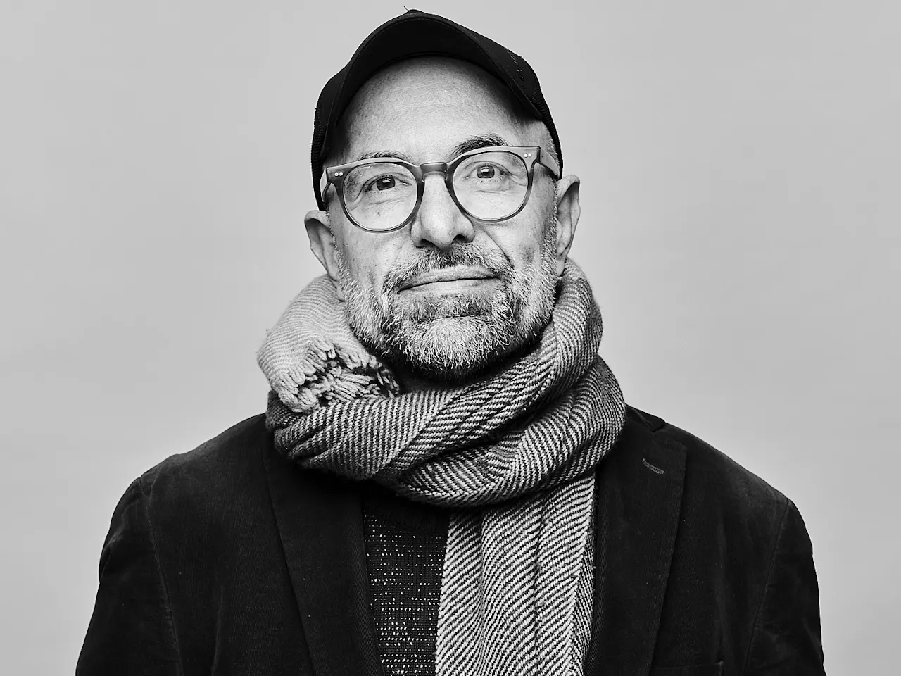 A black and white headshot of a person standing against a light wall wearing a black jacket, black hat, plastic rimmed glasses and a white knitted scarf. It is an image of the German director Philipp Stölzl.
