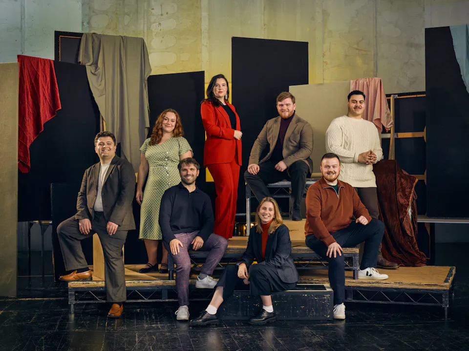 A group of people wearing different colour clothes pose either sitting or standing in a photographers studio which is decorated with draped partitions.