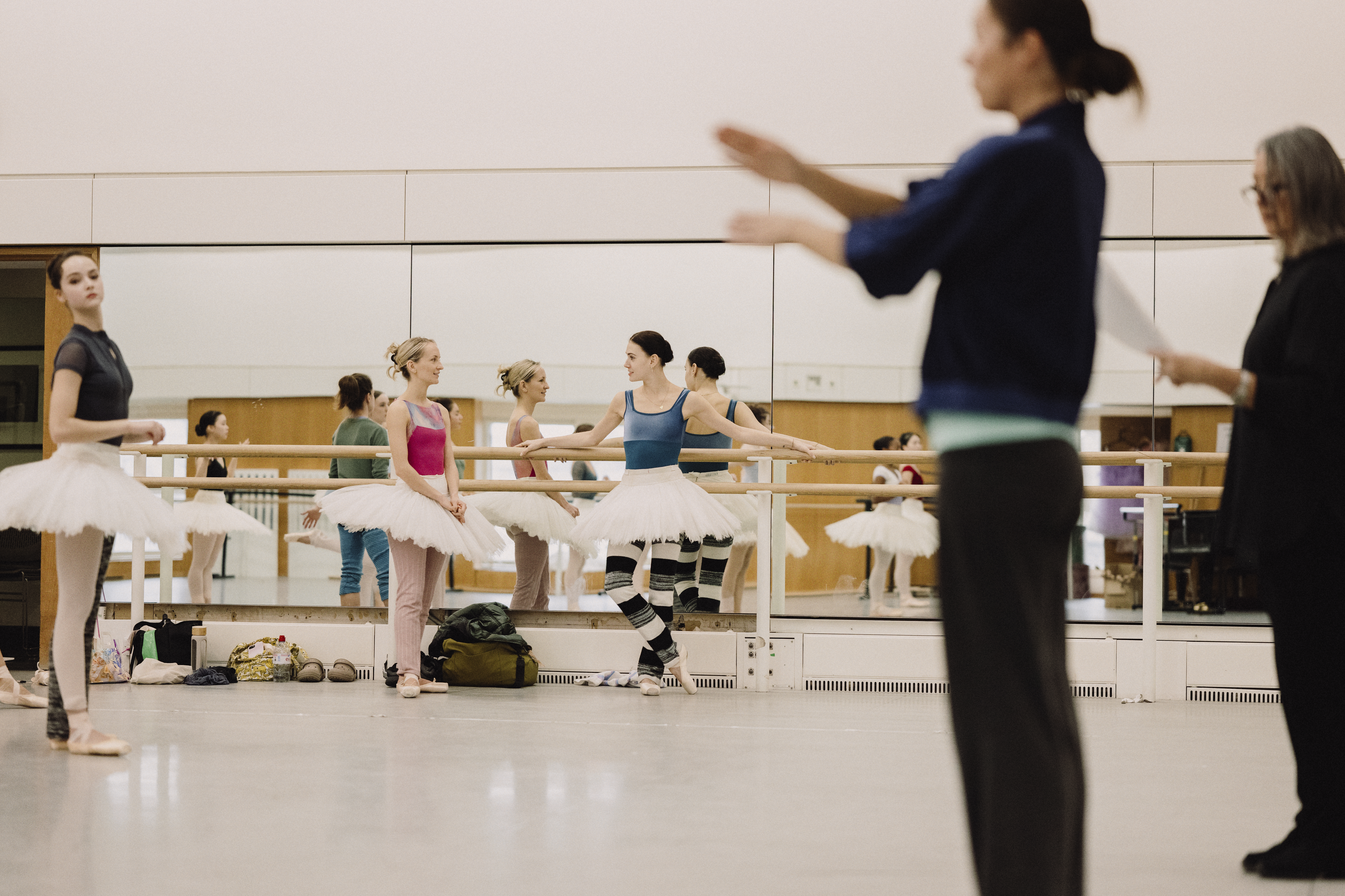 Several ballet dancers wearing tutu's stand in a brightly lit dance studio against a mirrored wall. In the corner two other people are coaching. 