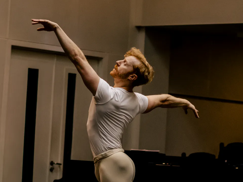 The Royal Ballet dancer Steven Mcrae stands in a well-lit studio with his arms stretched outwards.