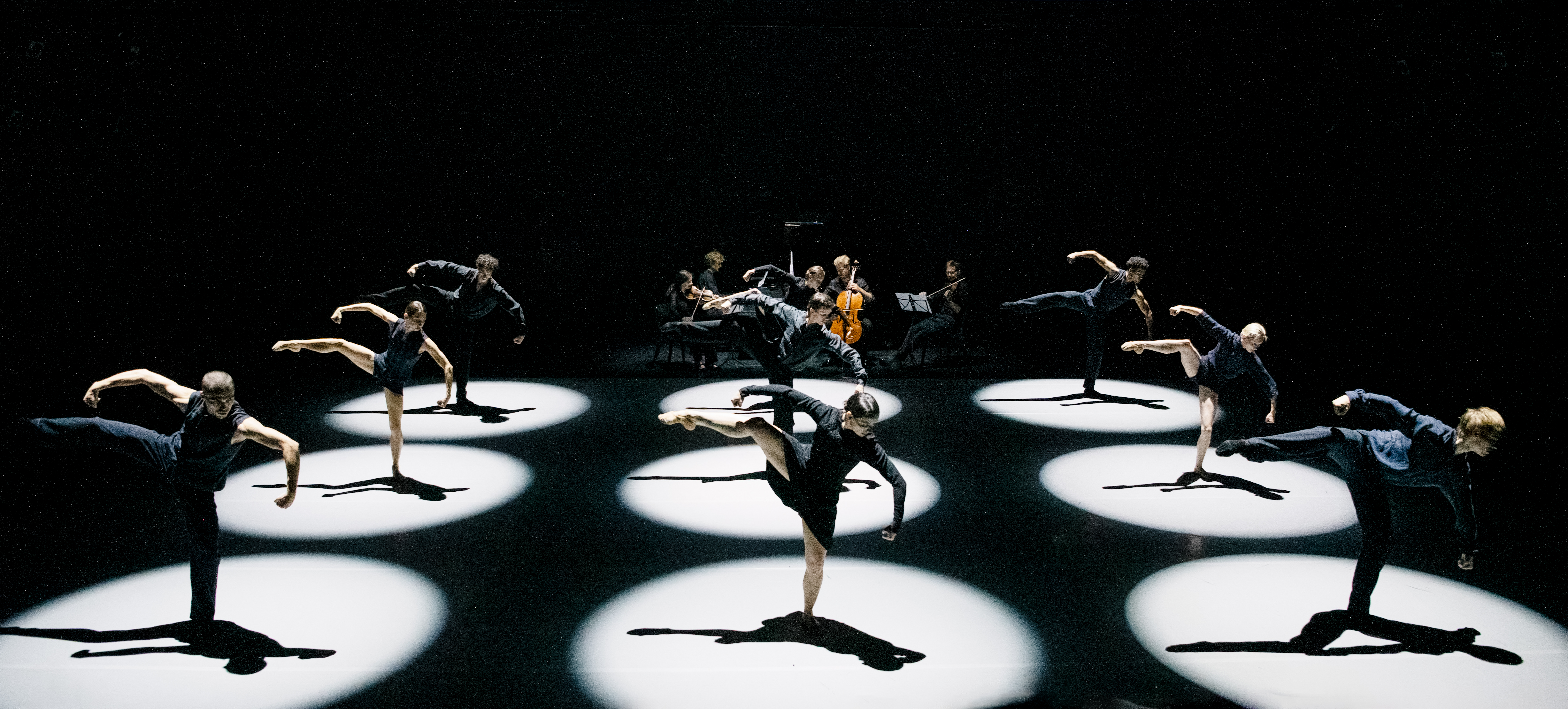 9 ballet dancers perform on stage within 9 brightly lit circular spotlights. All the dancers stand on one foot with their other stretched out. 