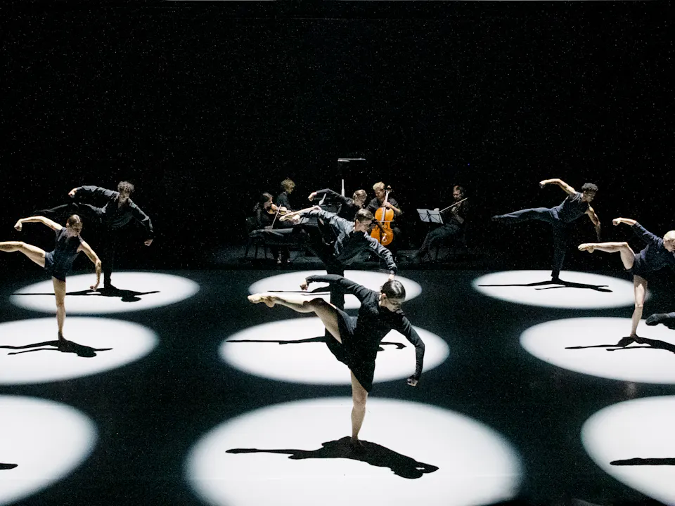 9 ballet dancers perform on stage within 9 brightly lit circular spotlights. All the dancers stand on one foot with their other stretched out.