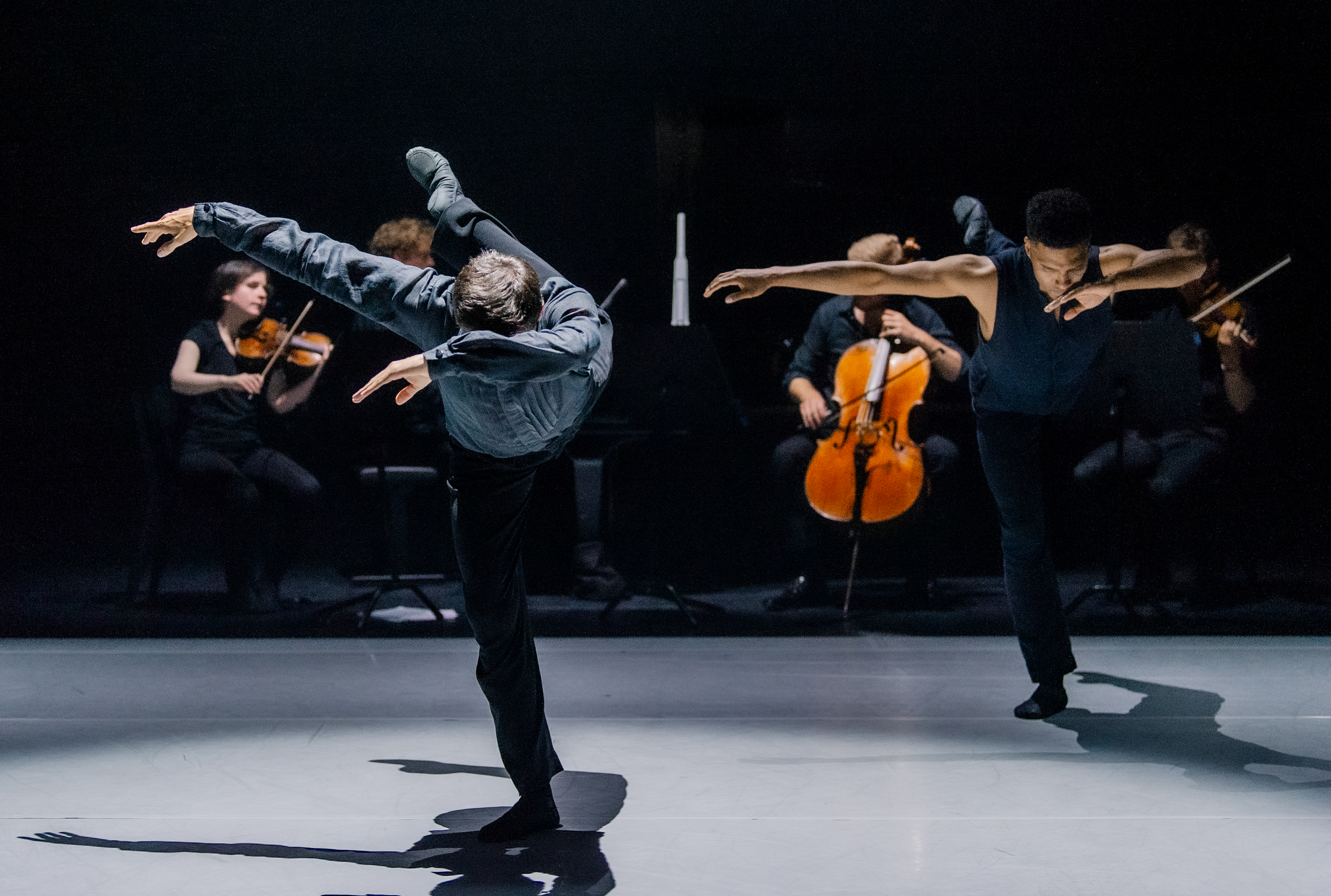 Two ballet dancers perform on a dark stage. Behind them, a few members of a string quartet are visible and performing. (3 of 4)