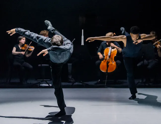Two ballet dancers perform on a dark stage. Behind them, a few members of a string quartet are visible and performing. (3 of 4)
