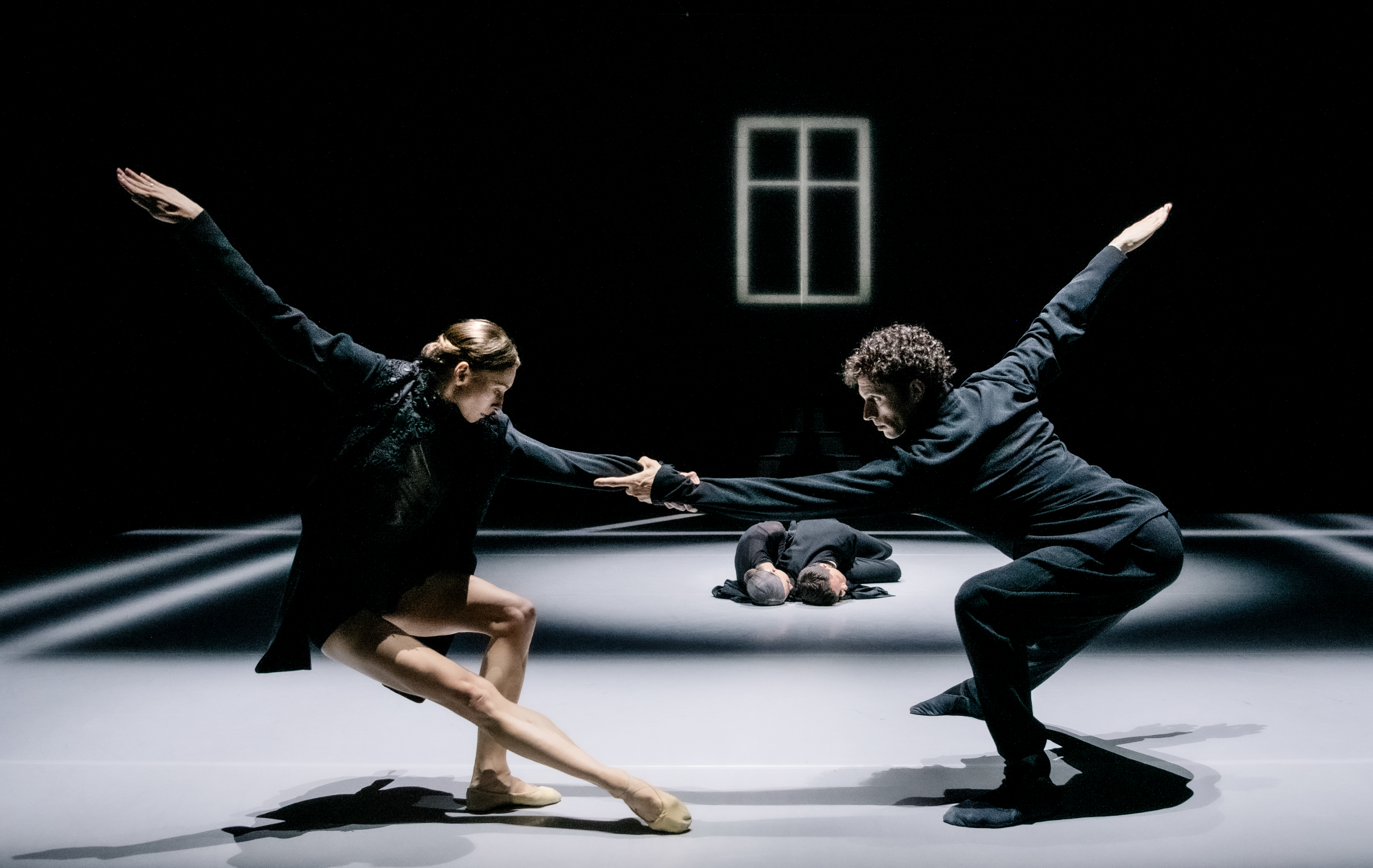 Two ballet dancers perform on a dark stage within a bright spotlight. In the background, two other dancers are lying on the floor embracing. (2 of 4)