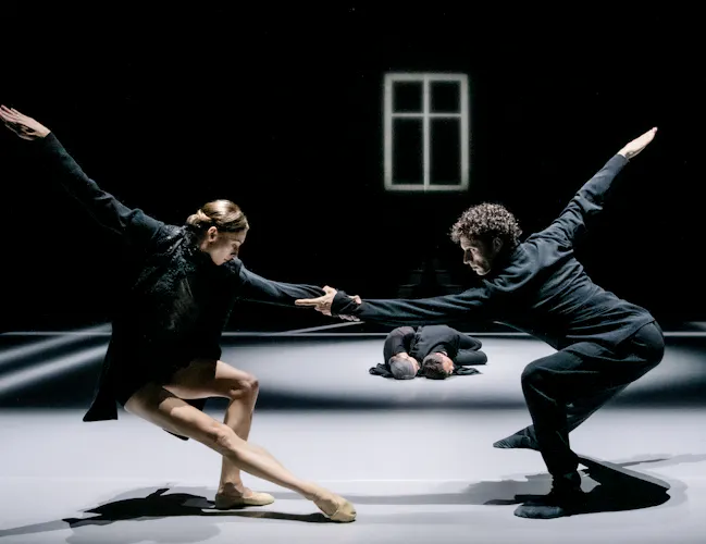 Two ballet dancers perform on a dark stage within a bright spotlight. In the background, two other dancers are lying on the floor embracing. (2 of 4)
