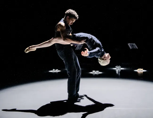 Two ballet dancers perform on a dark stage within a brightly lit circular spotlight. One dancer is upright holding the other by the waist as they drape backwards. (1 of 4)