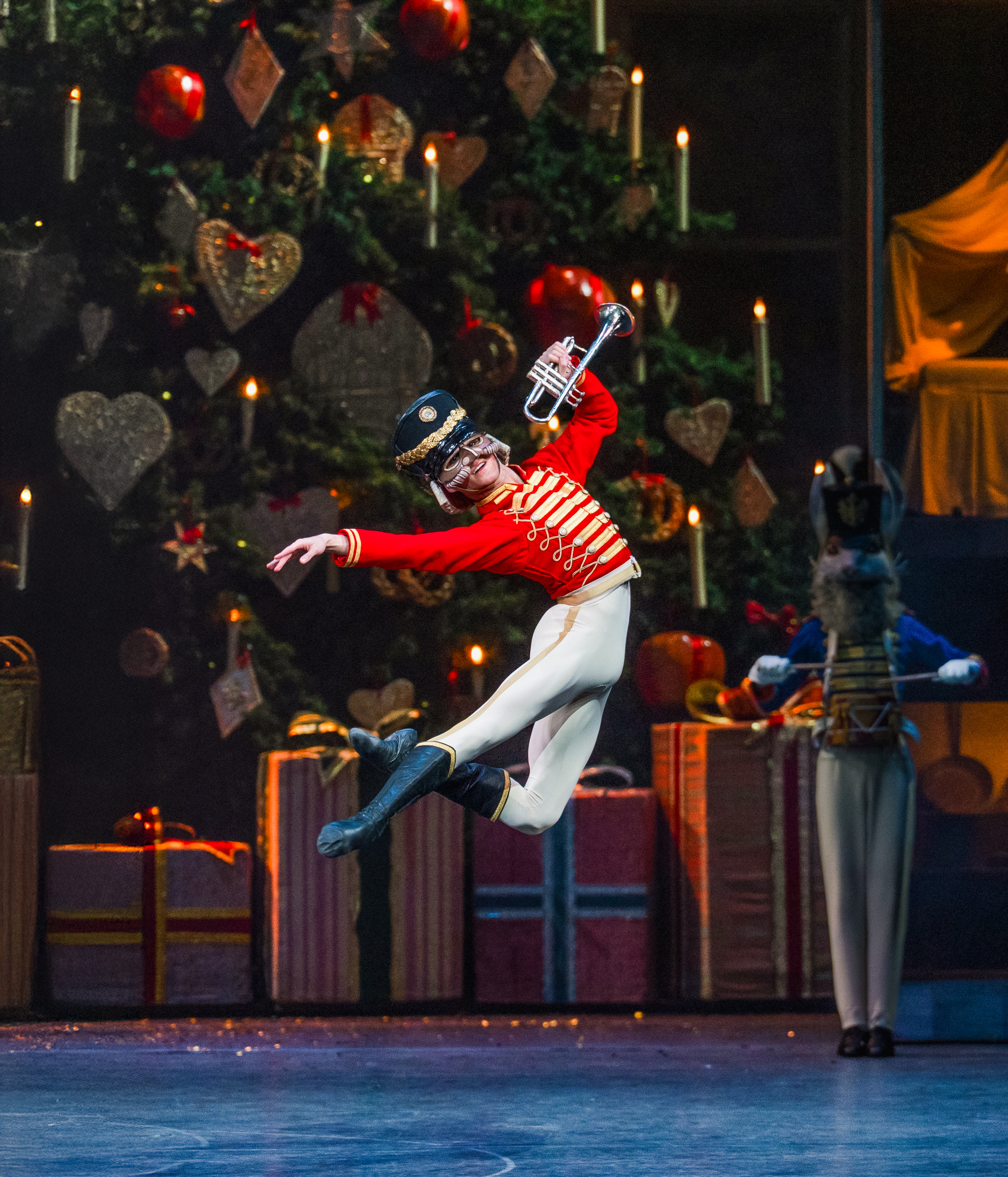 A man dressed in a red military jacket, white tights and black boots jumps in front of a large christmas tree with presents underneath.