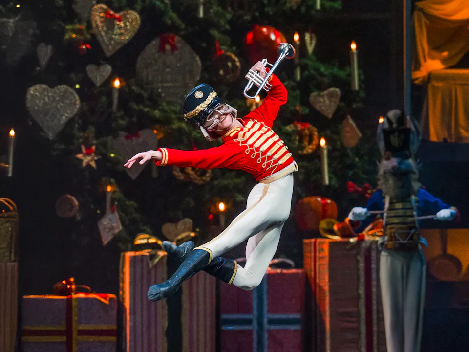 A man dressed in a red military jacket, white tights and black boots jumps in front of a large christmas tree with presents underneath.