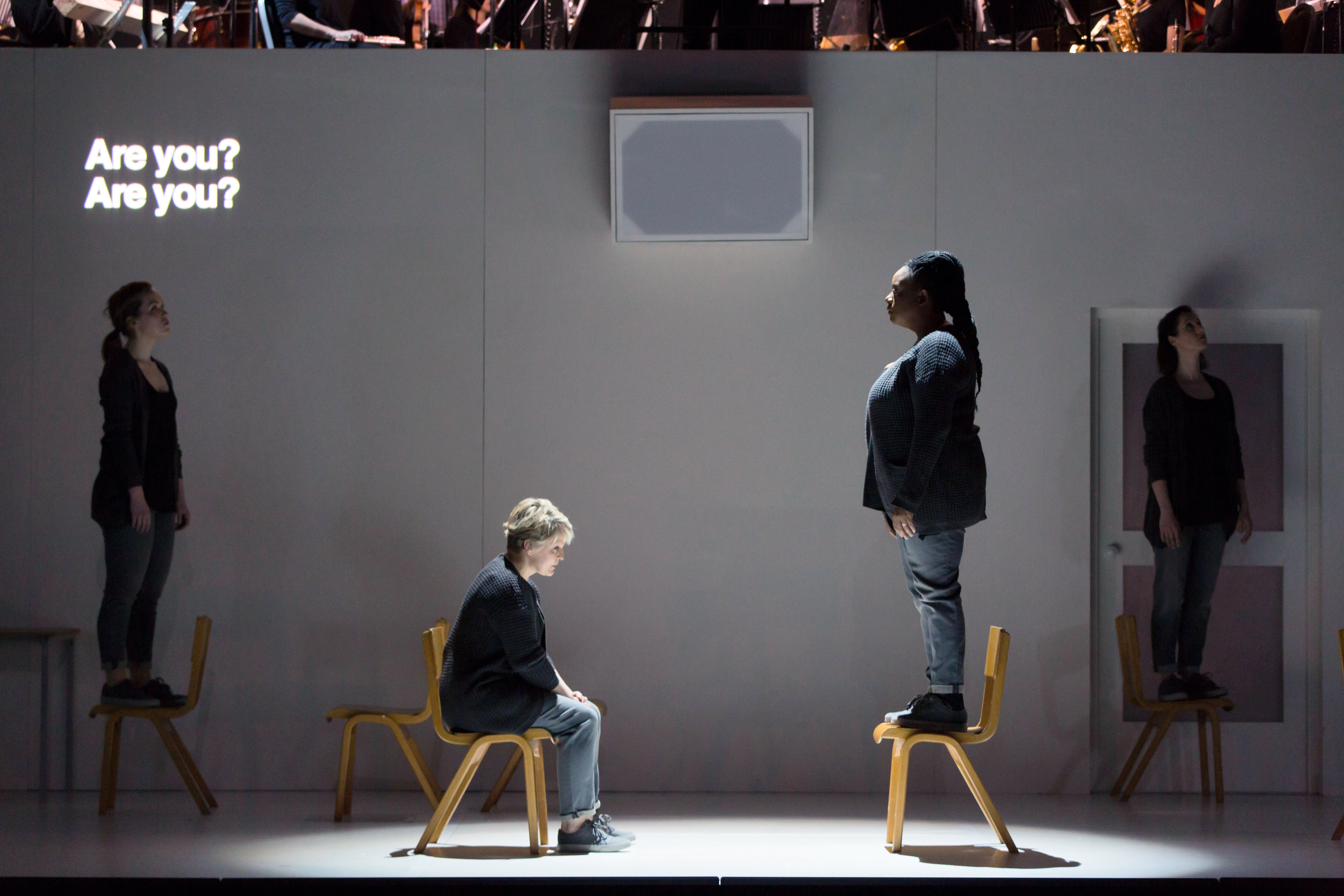A performer sits on a chair opposite another performer who is standing on the chair. They are in front of a white backdrop with the words "Are you? Are you" projected in the corner. Two other performers are in the foreground. (1 of 4)