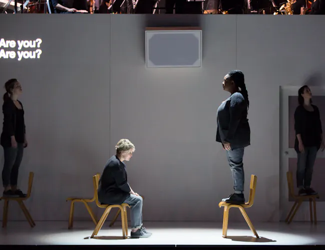 A performer sits on a chair opposite another performer who is standing on the chair. They are in front of a white backdrop with the words "Are you? Are you" projected in the corner. Two other performers are in the foreground. (1 of 4)