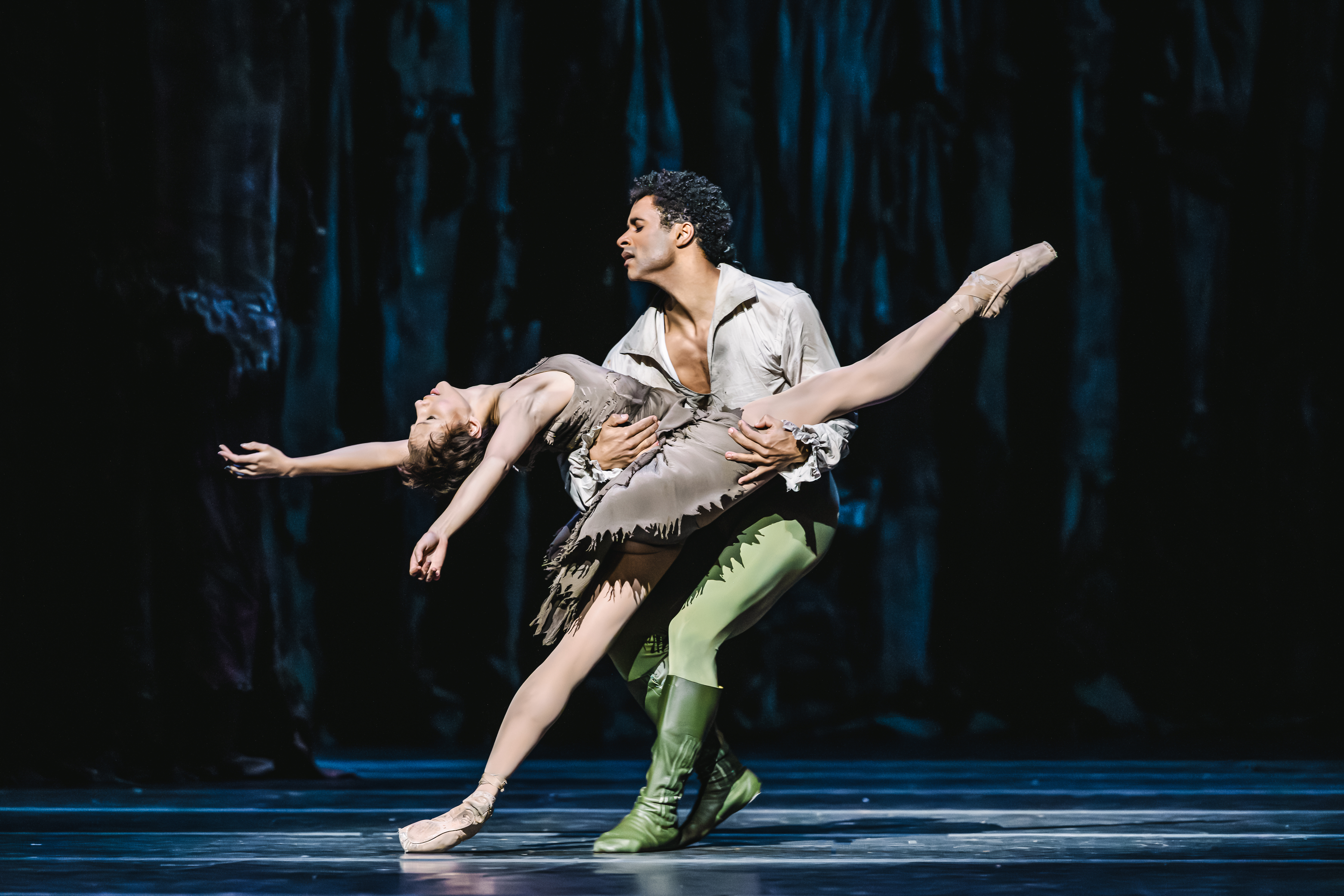 A male dancer wearing green tights and a brown shirt is holding a woman wearing a brown dress. She is leaning back with her leg outstretched across him, as he lunges near her.