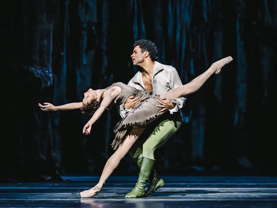 A male dancer wearing green tights and a brown shirt is holding a woman wearing a brown dress. She is leaning back with her leg outstretched across him, as he lunges near her.