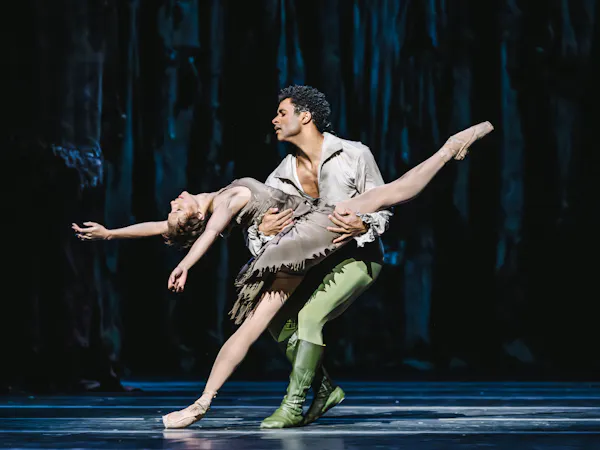 A male dancer wearing green tights and a brown shirt is holding a woman wearing a brown dress. She is leaning back with her leg outstretched across him, as he lunges near her.