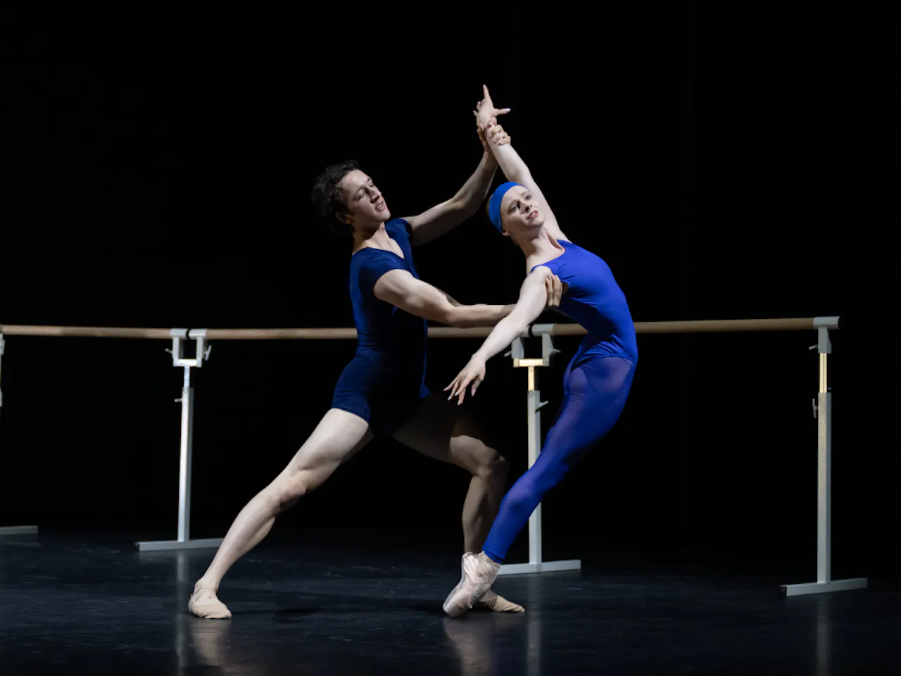 Two dancers in blue stand on a dark stage in front of a ballet barre. The woman leans her body forward with arms outstretched, she stands en pointe. Behind her the man holds her arms to support her.