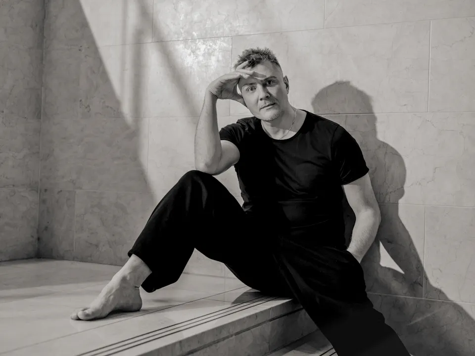 A black and white image of director Evgeny Titov, he wears black trousers and a black t-shirt and sits with his back against a concrete wall.