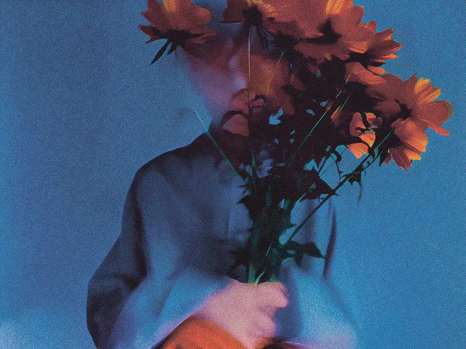 A stylized, grainy photograph of a person sitting against a blue-toned background, holding a bouquet of orange flowers that obscures their face. The person wears a muted blue top and a flowing orange skirt, with soft, moody lighting and a dreamy, slightly blurred effect.