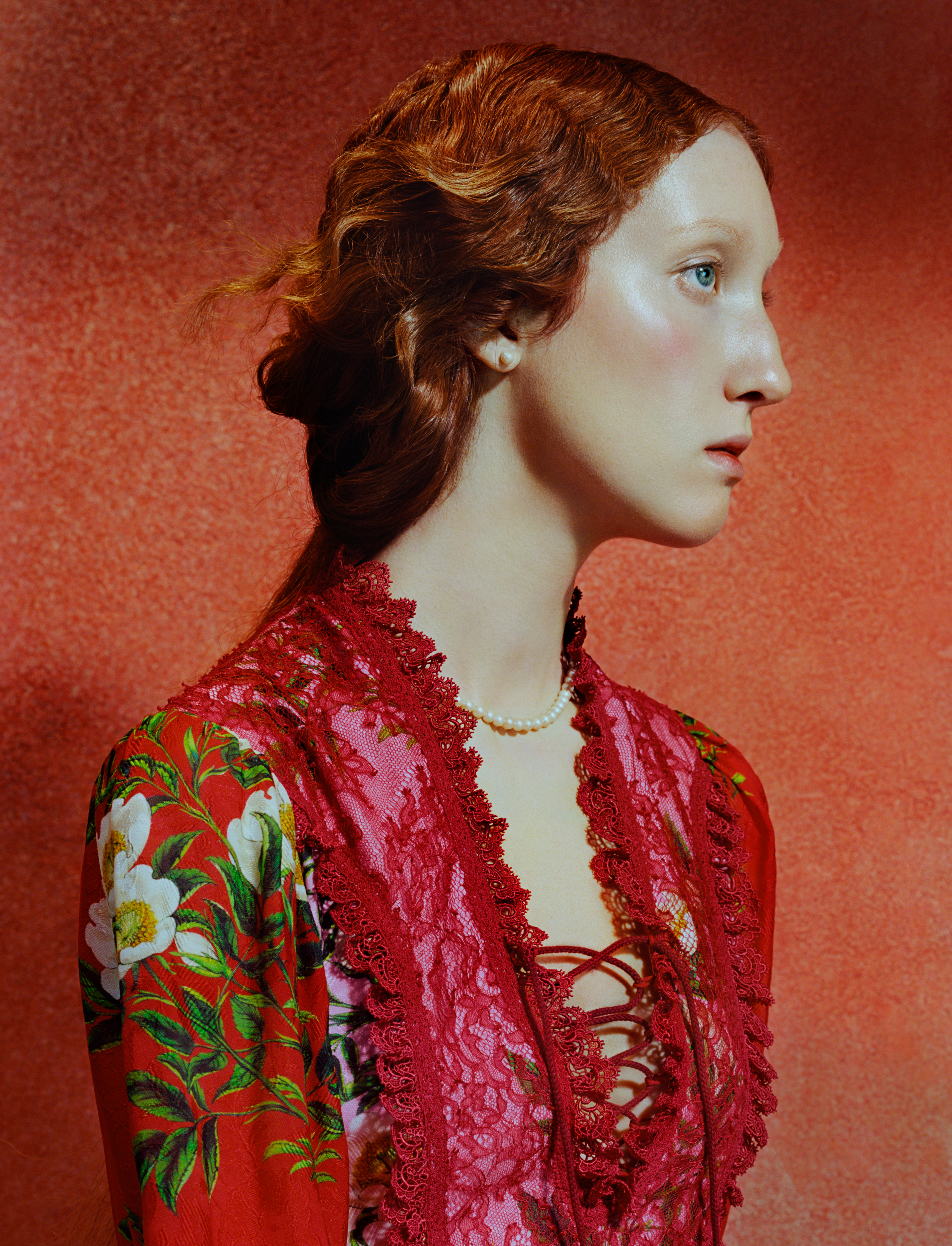 Close-up side profile of a young woman with pale skin and red hair styled in a loose updo, set against a warm red textured background. Soft lighting highlights her cheekbone and eye as she gazes forward with a calm, neutral expression.