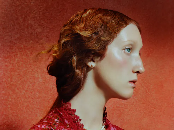 Close-up side profile of a young woman with pale skin and red hair styled in a loose updo, set against a warm red textured background. Soft lighting highlights her cheekbone and eye as she gazes forward with a calm, neutral expression.