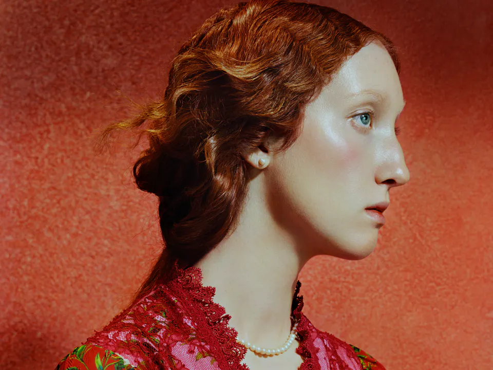 Close-up side profile of a young woman with pale skin and red hair styled in a loose updo, set against a warm red textured background. Soft lighting highlights her cheekbone and eye as she gazes forward with a calm, neutral expression.