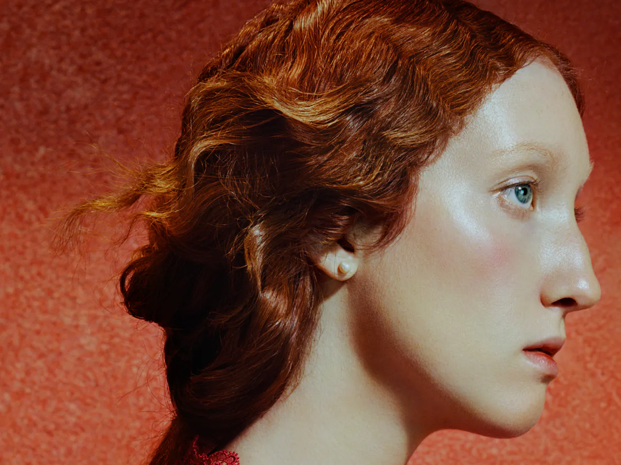 Close-up side profile of a young woman with pale skin and red hair styled in a loose updo, set against a warm red textured background. Soft lighting highlights her cheekbone and eye as she gazes forward with a calm, neutral expression.