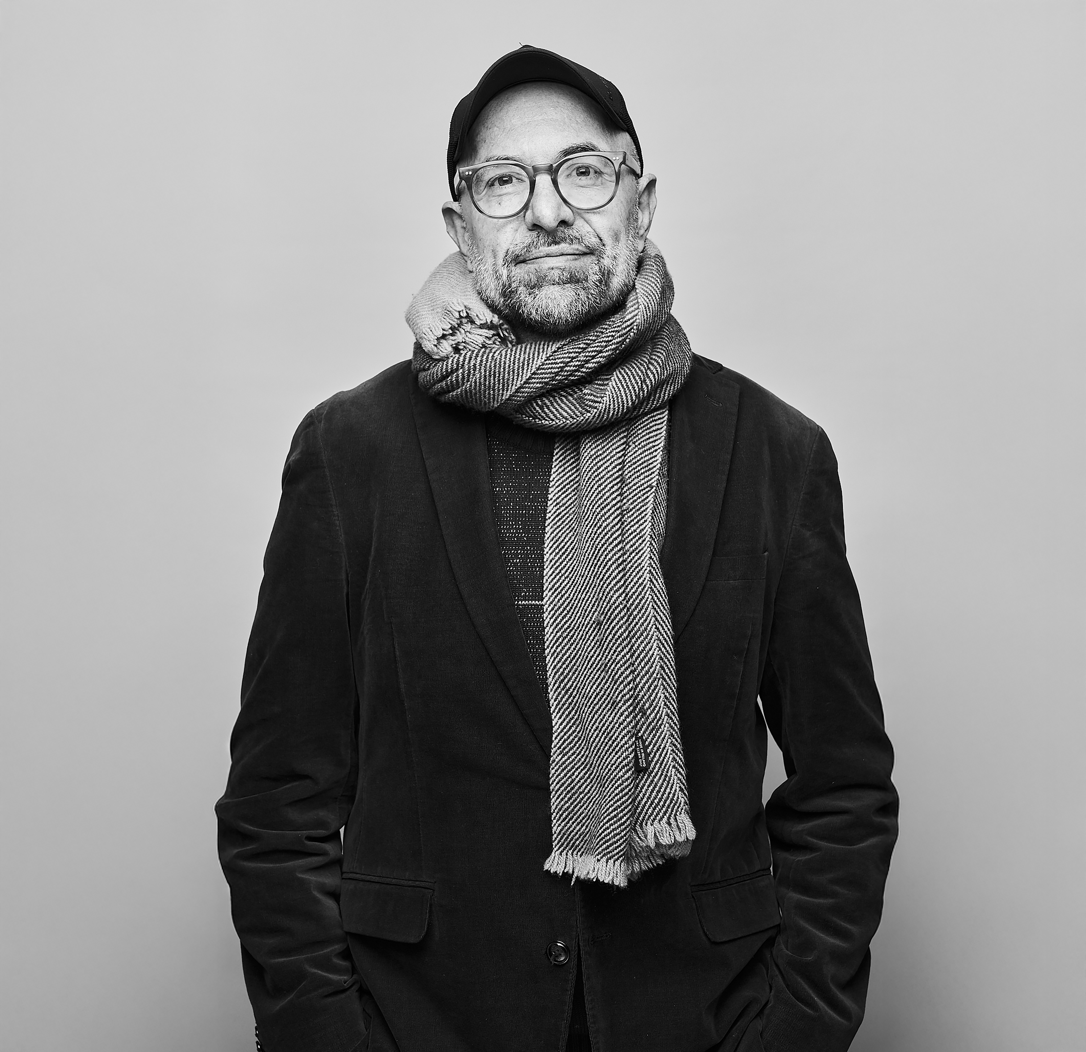 A black and white headshot of a person standing against a light wall wearing a black jacket, black hat, plastic rimmed glasses and a white knitted scarf. It is an image of the German director Philipp Stölzl.