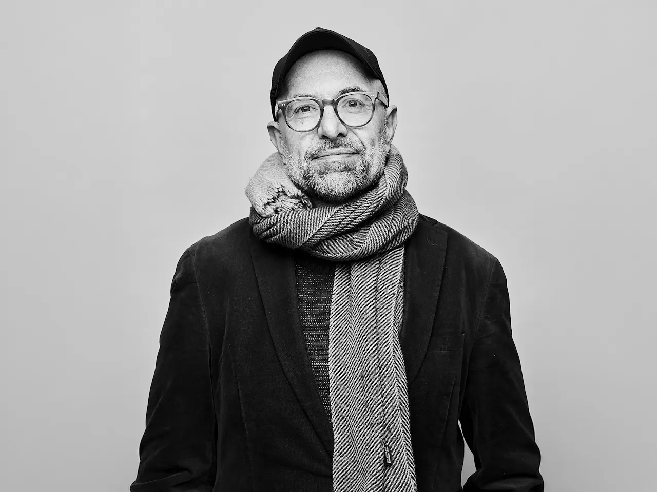 A black and white headshot of a person standing against a light wall wearing a black jacket, black hat, plastic rimmed glasses and a white knitted scarf. It is an image of the German director Philipp Stölzl.
