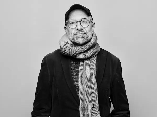 A black and white headshot of a person standing against a light wall wearing a black jacket, black hat, plastic rimmed glasses and a white knitted scarf. It is an image of the German director Philipp Stölzl.