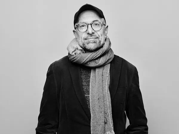 A black and white headshot of a person standing against a light wall wearing a black jacket, black hat, plastic rimmed glasses and a white knitted scarf. It is an image of the German director Philipp Stölzl.