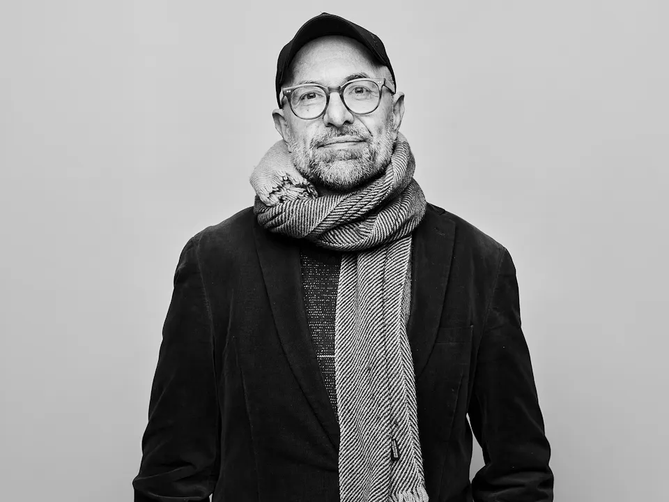A black and white headshot of a person standing against a light wall wearing a black jacket, black hat, plastic rimmed glasses and a white knitted scarf. It is an image of the German director Philipp Stölzl.