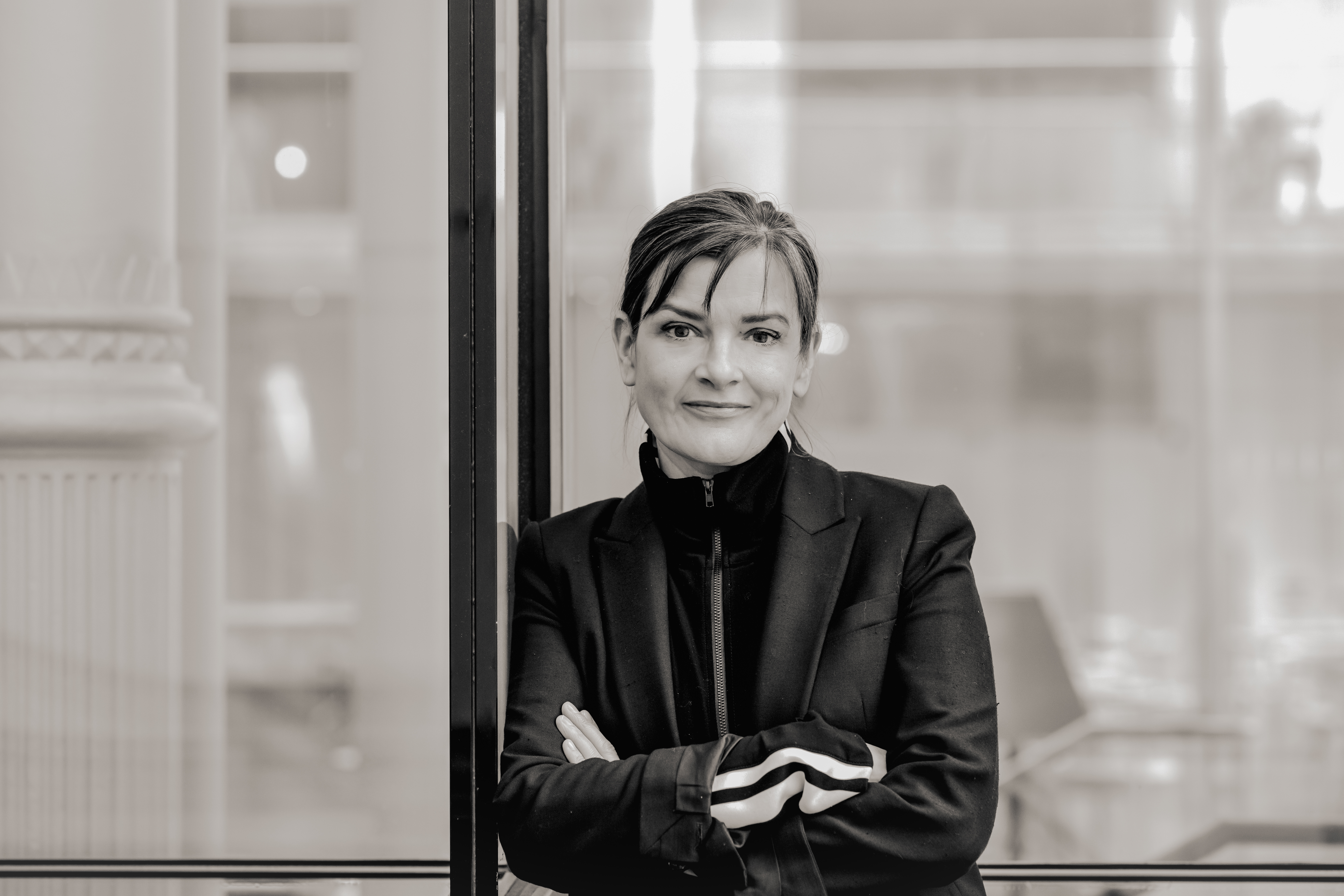 A black and white headshot of a person wearing a black jacket and black high necked jumper. It is a headshot of the Associate Director of The Royal Opera, Netia Jones. 